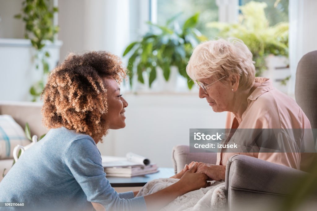 Cheerful home caregiver consoling senior woman Afro american female home nurse talking with senior woman, sitting in living room and holding her hands. Alzheimer's Disease Stock Photo