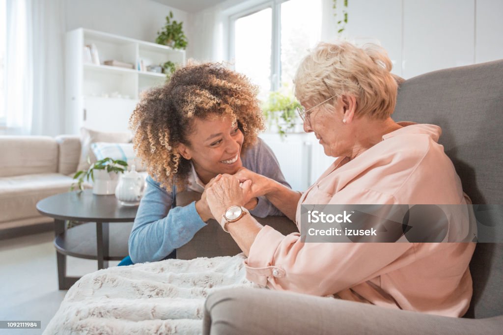 Cheerful nurse taking care of eldery lady Female home caregiver talking with senior woman, sitting in living room and holding hands. Alzheimer's Disease Stock Photo