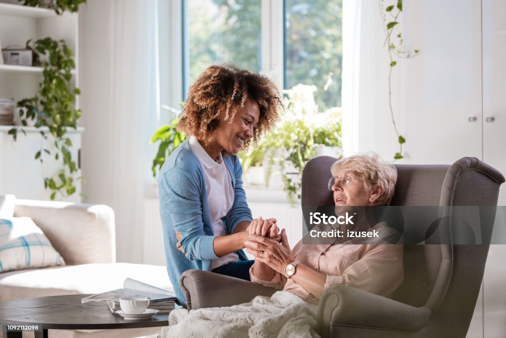 Cheerful nurse taking care of eldery lady Female afro american home caregiver taking care of senior woman, sitting in living room, listening to her and and holding her hands. Alzheimer's Disease Stock Photo