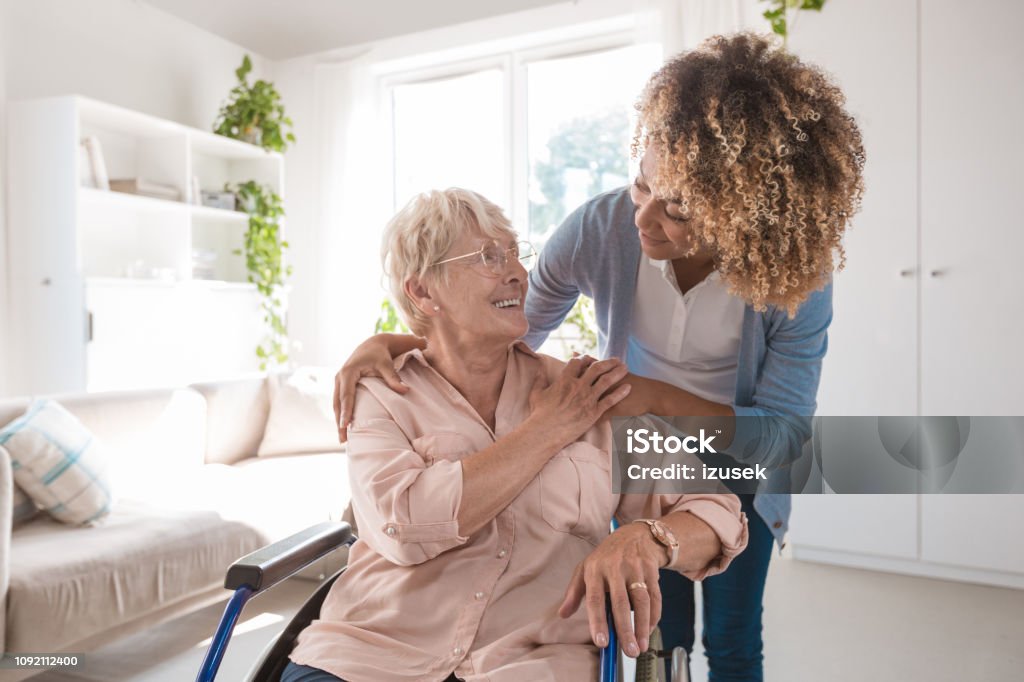 Cheerful nurse taking care of eldery lady Senior woman sitting in wheelchair and talking with afro american home caregiver, smiling to her. Senior Adult Stock Photo