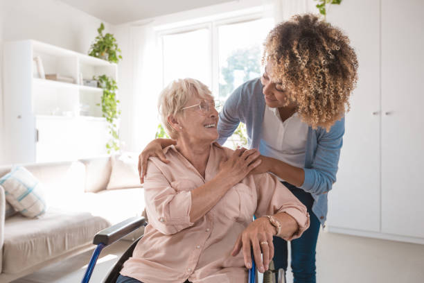 Cheerful nurse taking care of eldery lady Senior woman sitting in wheelchair and talking with afro american home caregiver, smiling to her. parkinsons disease photos stock pictures, royalty-free photos & images