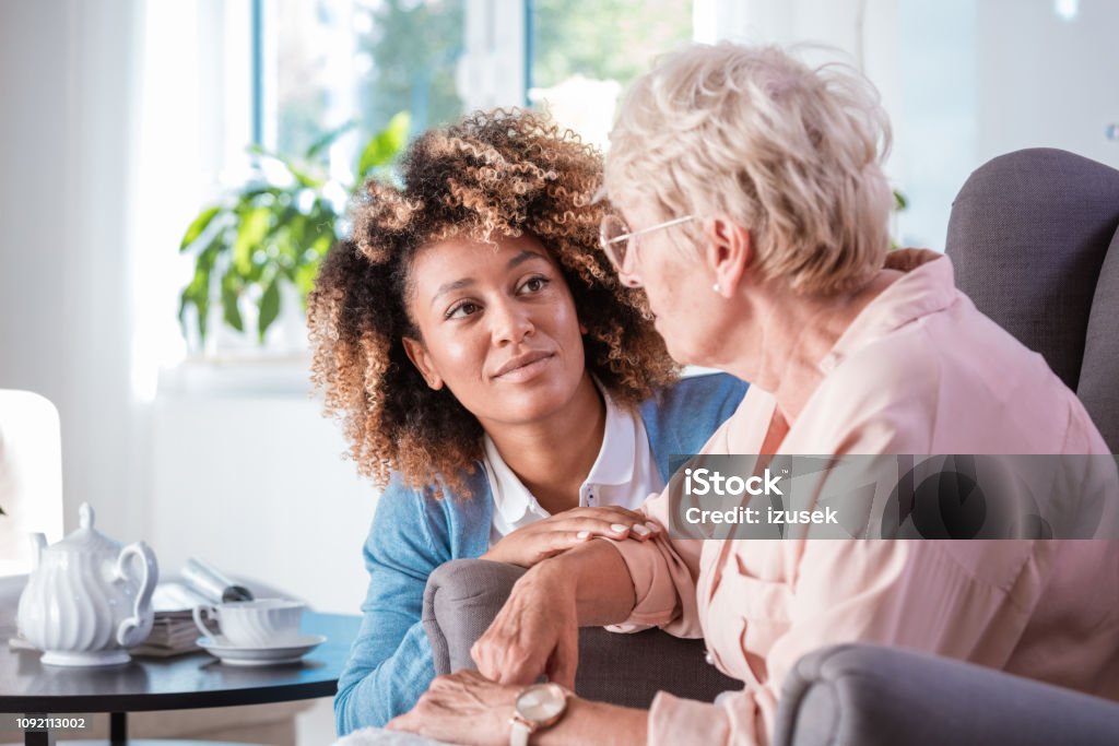 Friendly nurse taking care of eldery lady Female home caregiver talking with senior woman, sitting in living room and listening to her carefully. Alzheimer's Disease Stock Photo
