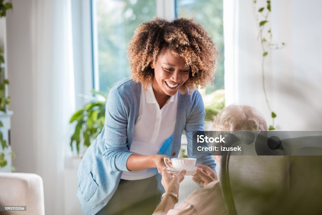 Home caregiver serving tea to senior woman Cheerful afro american female home caregiver supporting senior woman, serving her tea. Community Outreach Stock Photo