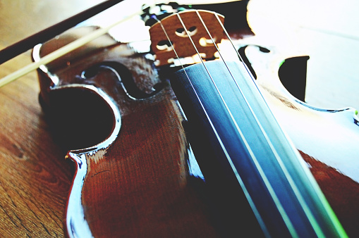 A violin artistically shot laterally, partially visible, over a wooden surface. The upper and lower bout, bouts, the waist, the F holes, The strings and the bow are visible in the frame.There is a white light from top right corner falling on the violin and making it bright and aesthetic. Depth of field. There is beautiful wooden pattern marks over the surface.