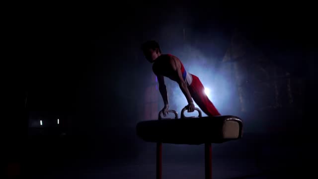 male gymnast athlete performs handstand and spin on Pommel horse on a dark background and smoke in slow motion. Olympic programme. Preparation for the exercise
