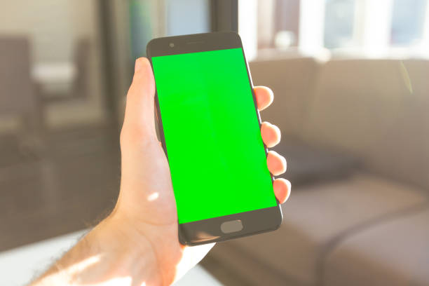 Male hand holding smartphone with green screen indoors sunlight stock photo