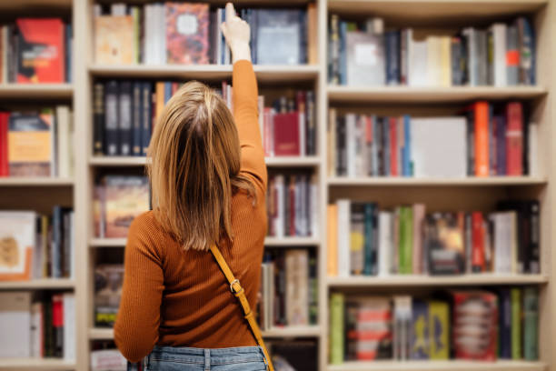 Young girl student Pretty young girl with loose long blondie hair standing and holding an open book between book shelves in the library, reading and looking into the book bookstore stock pictures, royalty-free photos & images