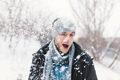 Man portrait being the reached target in a snow fight. Snowflakes covering his face, closed eyes, smile, having fun. Winter entertainment outdoors. Copy space.