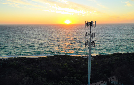 Aerial view of a cell phone tower (mobile phone tower) at sunset, with the orange sun setting above a blue ocean and clouds in the sky