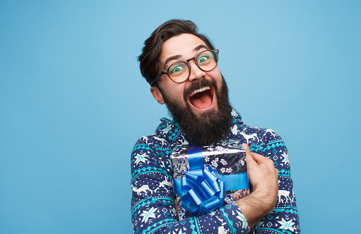 Excited bearded guy in eyeglasses and sweater embracing packed present and looking at camera on blue background.
