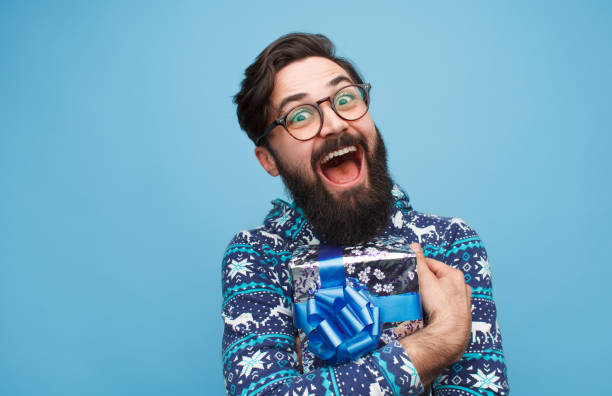 Crazy man happy with present Excited bearded guy in eyeglasses and sweater embracing packed present and looking at camera on blue background. christmas excitement stock pictures, royalty-free photos & images