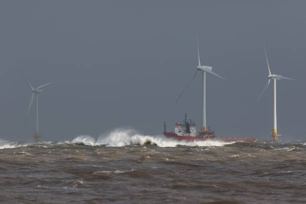 Ship sailing in rough sea around offshore wind farm turbines. Ship sailing in rough sea. High cost energy of offshore wind farm turbines. Supply vessel battling ocean swell. Dangerous waves for shipping in storm conditions. Grey sky background with copy space offshore wind farm stock pictures, royalty-free photos & images