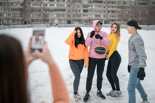 A group of young persons dressed in a casual clothing are going to play basketball outdoors at winter. The young woman is holding the mobile phone and taking the picture of the friends. The young man is pumping the ball. Shooting at winter day in a courtyard of a residential building