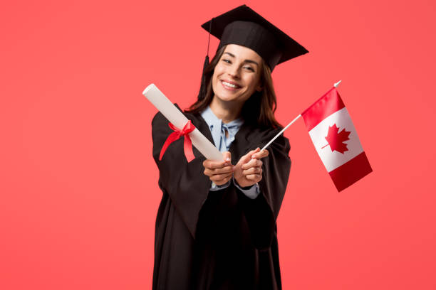 smiling female student in academic gown holding canadian flag isolated on living coral - canadian flag fotos imagens e fotografias de stock