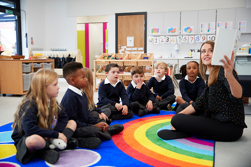 Maestra, sosteniendo un libro frente a su clase de la escuela primaria los niños sentados en el suelo en un salón, vista lateral photo