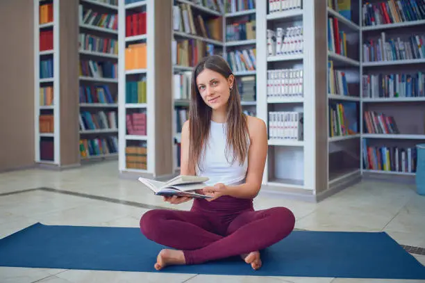 Beautiful young woman reading book and practices yoga asana Sukhasana - The Easy Sitting crosslegged Pose in the library.