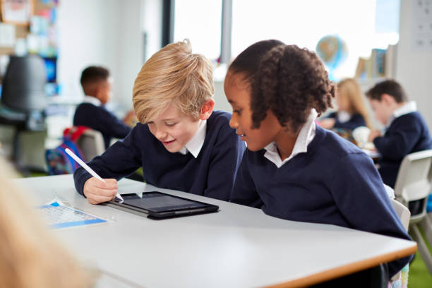 Two primary school kids sitting together at desk in a classroom using a tablet computer and stylus, close up Two primary school kids sitting together at desk in a classroom using a tablet computer and stylus, close up school children stock pictures, royalty-free photos & images
