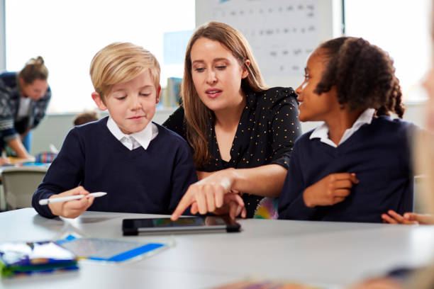 istock Female school teacher helping two kids using a tablet computer at desk in a primary school classroom, front view 1092193620