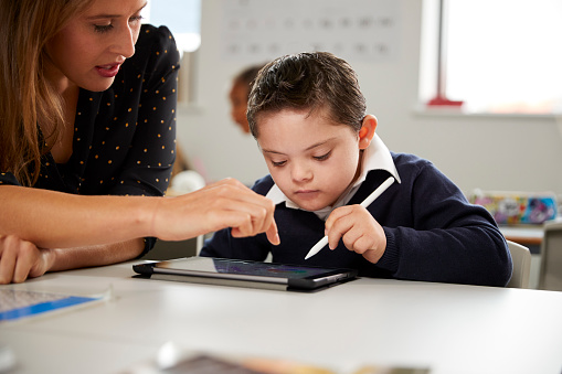 Joven maestra trabajo con un niño síndrome de Down sentado en el escritorio utilizando un equipo tablet PC en un aula de escuela primaria, vista frontal, primer plano photo