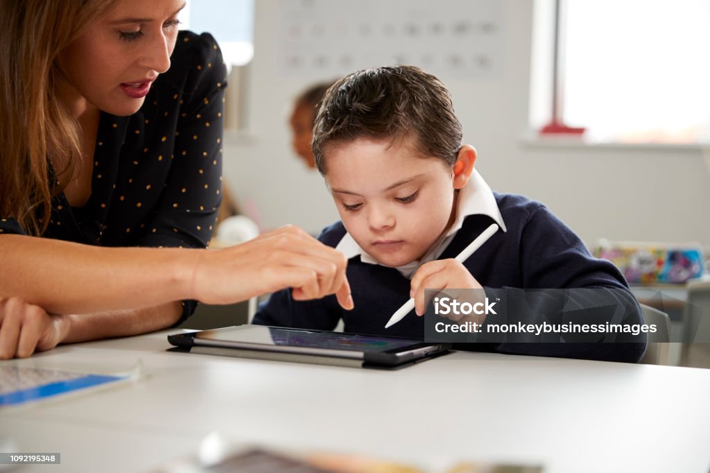 Young female teacher working with a Down syndrome schoolboy sitting at desk using a tablet computer in a primary school classroom, front view, close up Child Stock Photo