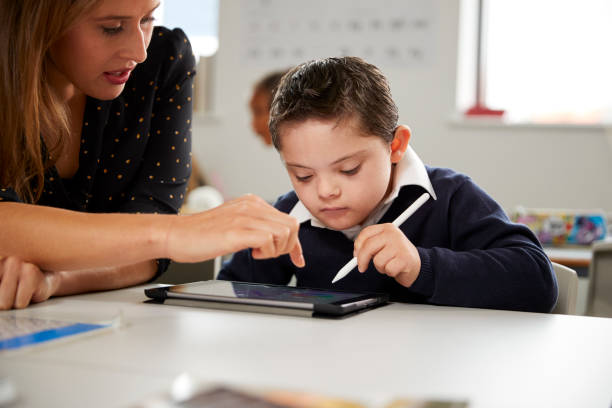 Young female teacher working with a Down syndrome schoolboy sitting at desk using a tablet computer in a primary school classroom, front view, close up Young female teacher working with a Down syndrome schoolboy sitting at desk using a tablet computer in a primary school classroom, front view, close up special education stock pictures, royalty-free photos & images