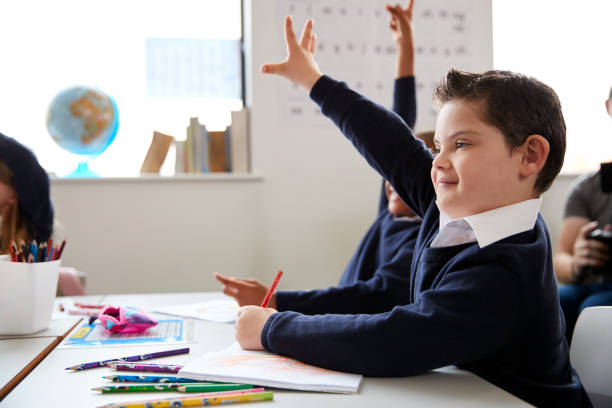 Schoolboy with Down syndrome sitting at a desk raising his hand in a primary school class, close up, side view Schoolboy with Down syndrome sitting at a desk raising his hand in a primary school class, close up, side view special education stock pictures, royalty-free photos & images