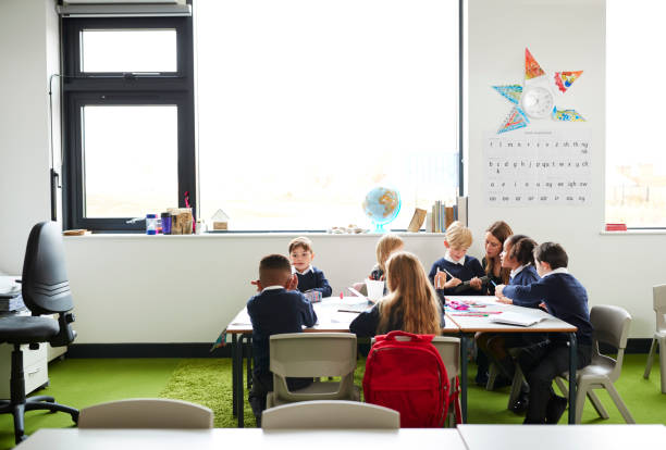 istock A group of primary school kids sitting at table in a classroom, teacher kneeling to help them during the lesson 1092197302