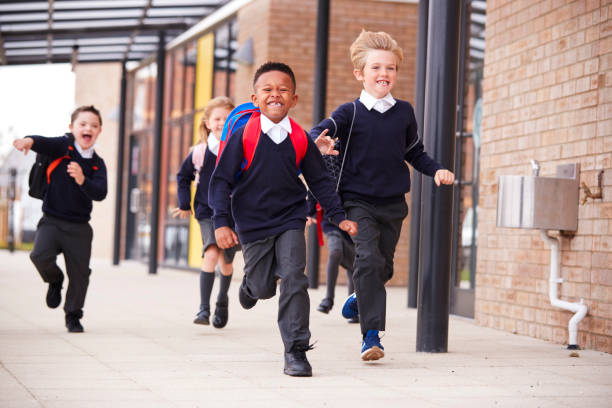 cerca de escuela primaria feliz niños, vistiendo uniformes y mochilas, corriendo en una calzada fuera de su edificio de escuela, vista frontal, - uniforme de colegio fotografías e imágenes de stock