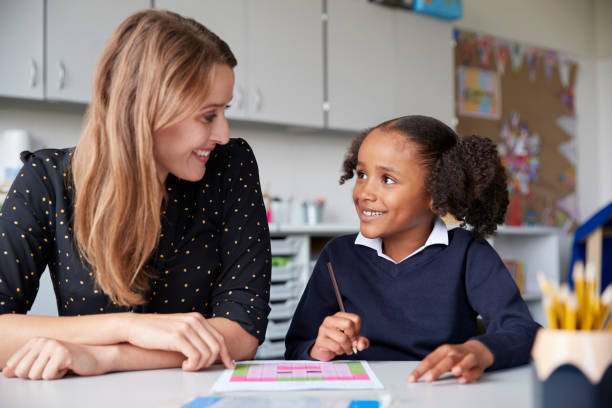 giovane insegnante di scuola elementare che lavora una contro una con una studentessa a un tavolo in classe, entrambe che si guardano sorridenti, da vicino - one on one foto e immagini stock