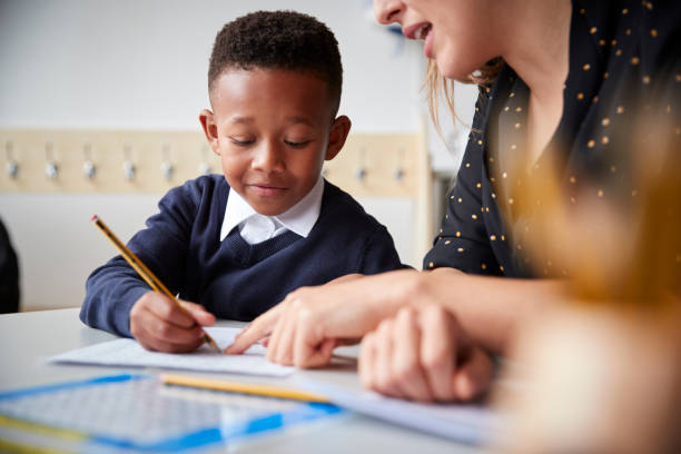 female primary school teacher helping a young school boy sitting at table in a classroom, close up, selective focus - teachers school student imagens e fotografias de stock