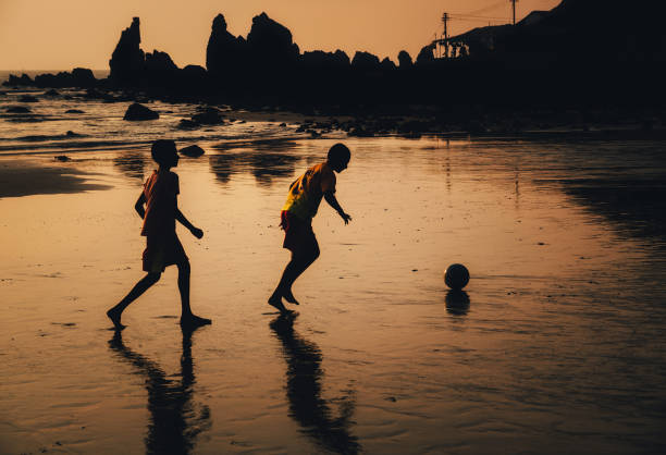 Arambol, Goa, India - December 08, 2014: Silhouettes of two boys play soccer or football on the indian beach at sunset against the cliffs, North Goa. Lifestyle, vacation, happiness, joy and carefree childhood concept