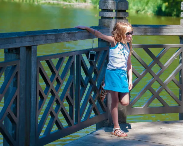 A little girl leaning against a railing of a deck along a pond at a park resting after taking pictures with her camera.