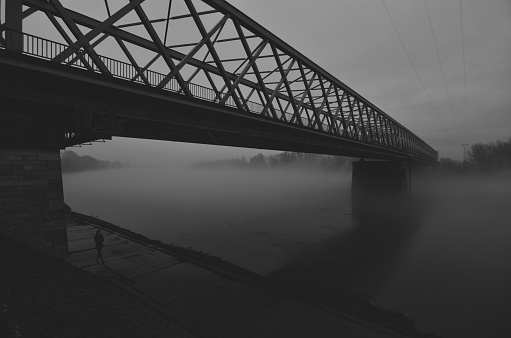 Silhouette of the young woman in black clothes standing alone under the bridge on a misty autumn day.