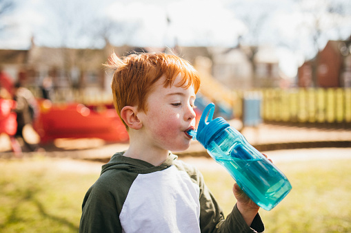 Little boy drinking from a blue plastic water bottle in the park.