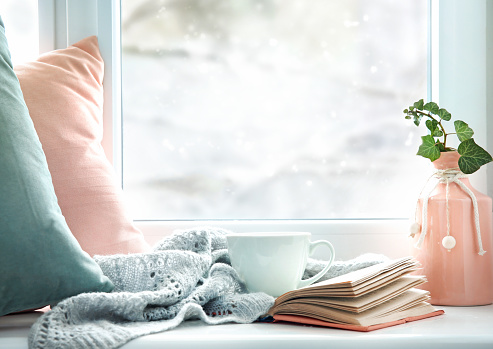 Winter life style background.Mug of tea, book,pillows on window sill empty copy space backdrop.Cold weather mood concept backdrop.