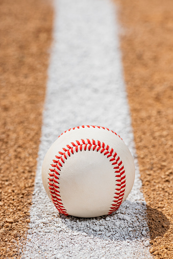 A low view of a new baseball sitting on a painted white Foul Line on dirt of a baseball diamond infield. Sometimes this can be referred to as a Fair Line as it is in Fair Territory and the area from the edge of the line is Foul Territory.