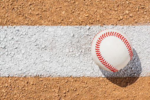 An overhead view of a new baseball sitting on a painted white Foul Line on dirt of a baseball infield diamond. Sometimes this can be referred to as a Fair Line as it is in Fair Territory and the area from the edge of the line is Foul Territory.