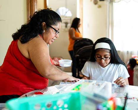 The mother, body-positive Mexican woman, teaching her teenage daughter how to do a craft, fancy beadworks. They are sitting around the big round table in the living room. Pennsylvania, USA.