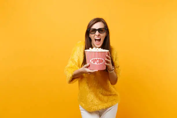 Crazy young girl in 3d imax glasses watching movie film screaming holding bucket of popcorn isolated on bright yellow background. People sincere emotions in cinema lifestyle concept. Advertising area