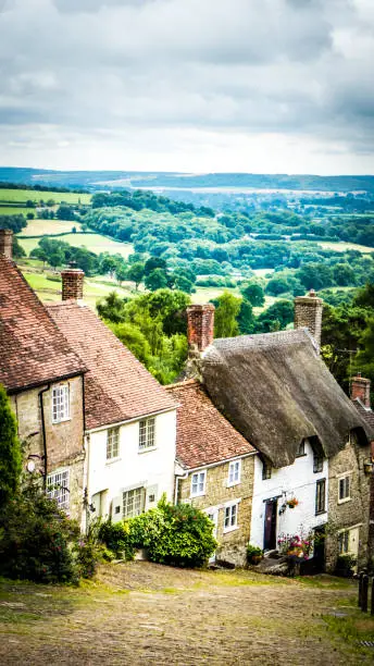 Gold Hill in Shaftesbury is an authentic place in the South of the UK. View of a stone old road on a hill with old English limestone houses with thatched roofs in the background in Dorset countryside.