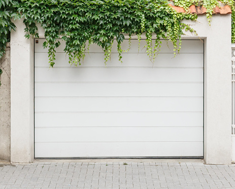 White closed wooden single garage doors in front covered with green ivy foliage. Grey tiles near.