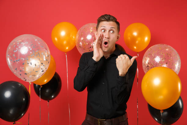 istock Amazed young man in black classic shirt looking aside, pointing thumb aside, keeping hand on cheek on red background air balloons. Valentine's Day Happy New Year birthday mockup holiday party concept. 1092355990