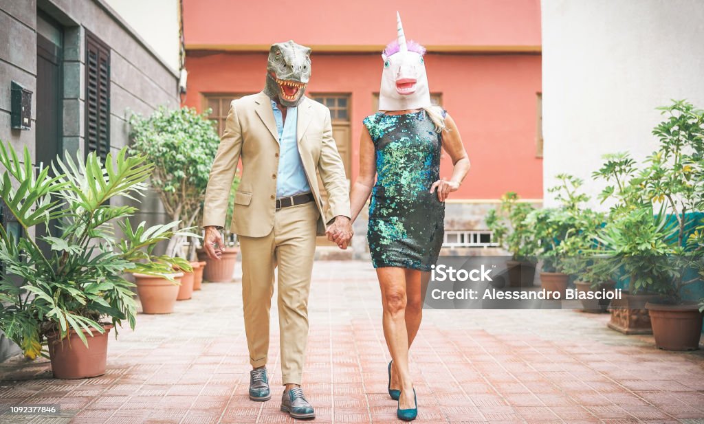 Crazy senior couple wearing dinosaur and unicorn mask - Mature trendy people having fun masked at carnival parade - Absurd, eccentric, surreal, fest and funny masquerade concept Costume Stock Photo