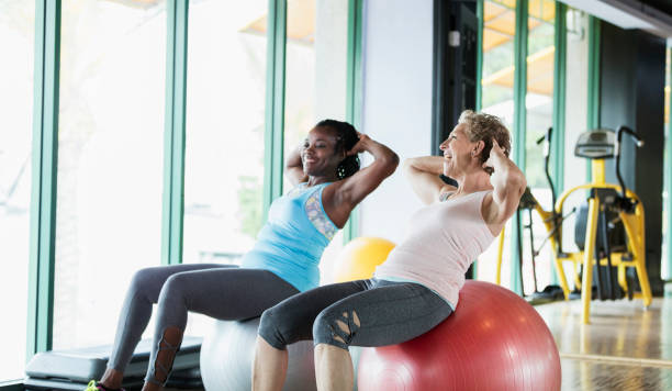 Two women at the gym on medicine balls, talking Two multi-ethnic women exercising together at the gym, sitting side by side on giant medicine balls, hands behind their heads. The senior woman, in her 60s, is talking to her African-American friend, a mature woman in her 50s. fitness ball photos stock pictures, royalty-free photos & images
