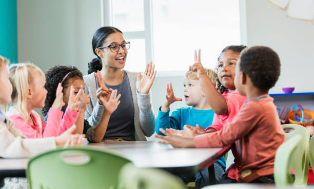 Multi-ethnic preschool teacher and students in classroom A multi-ethnic group of six preschool children with a mixed race African-American and Caucasian teacher, sitting around a table in a classroom. The teacher and some of her students have their hands raised, holding up fingers, learning how to count. The children are 4 years old. nursery school child stock pictures, royalty-free photos & images