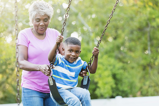 A senior African-American woman in her 60s with her 4 year old grandson at the park. She is pushing him on a swing on the playground. He is smiling at the camera.