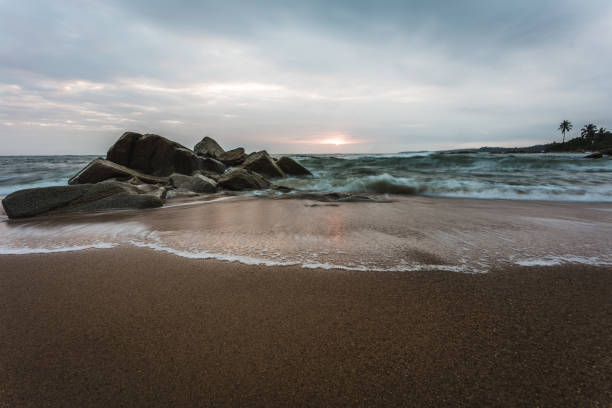 A rocky beach with white smooth waves from the blue ocean and a dramatic cloudy sky during a moody sunrise with deep blue and orange colors in Ghana, Africa