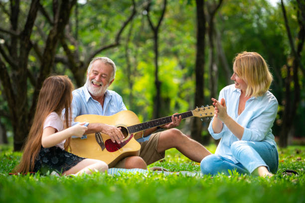 Happy family play guitar and sing together in park Happy family play guitar and sing together while sitting in the park in summer. Concept of family bonding by music. dv stock pictures, royalty-free photos & images