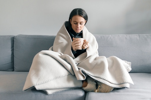 Autumn winter portrait of young girl resting at home on the sofa with cup of hot drink, under warm blanket.