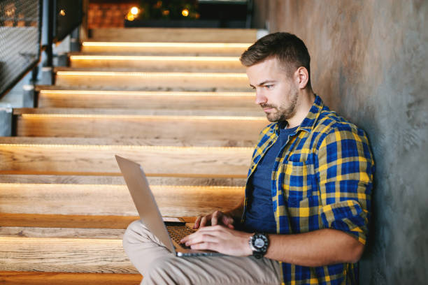 istock Serious bearded handsome freelancer in shirt using laptop for work while sitting on stairs indoors. Hands on keyboard. 1092547484